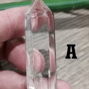 Può includere: Una punta di cristallo di quarzo trasparente con un design sfaccettato. Il cristallo è tenuto in mano, con la lettera "A" in nero a destra. Il cristallo è lungo circa 7.6 cm.