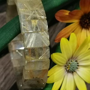 May include: A clear, rectangular bracelet with golden inclusions. The bracelet is made of multiple rectangular pieces. The background includes green stems and yellow and orange flowers.
