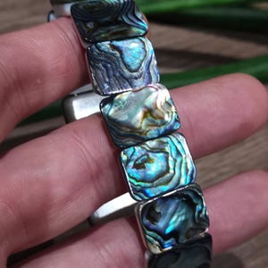 Può includere: Primo piano di un braccialetto con perline quadrate di conchiglia di abalone. Le perline iridescenti mostrano blu, verdi e viola, con una montatura in metallo argentato. Il braccialetto è mostrato su uno sfondo sfocato.