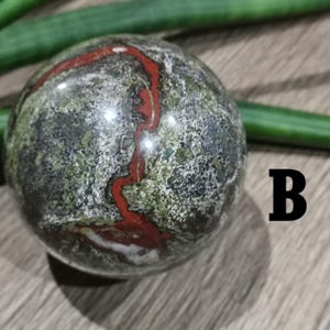Könnte beinhalten: Eine grüne, schwarze und rote Kugel mit einem wirbelnden Muster. Die Kugel ist aus poliertem Stein.