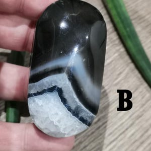 Puede incluir: Una piedra pulida de forma ovalada con un llamativo patrón de bandas negras, blancas y azules. La piedra tiene un acabado brillante y una base cristalina texturizada. La letra "B" es visible en el lado derecho de la piedra.
