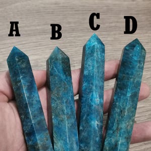 Peut inclure: Quatre pointes de cristal d'apatite bleue, étiquetées A, B, C et D, sont tenues dans une main. Les pointes sont toutes à peu près de la même taille et de la même forme, et elles ont une finition lisse et polie.