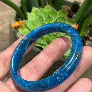 May include: A polished, deep blue bangle bracelet with a smooth, rounded surface. The bangle displays natural variations in colour and texture throughout its circular form, showcasing a vibrant, translucent blue appearance with subtle internal inclusions.