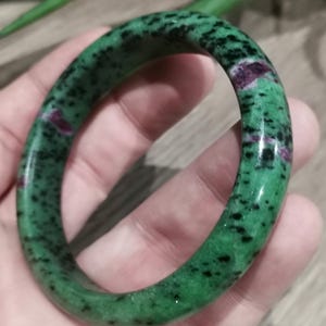 Peut inclure: Un bracelet en pierre précieuse vert et noir moucheté. Le bracelet a une surface lisse et polie avec un mélange de couleurs vertes et noires. Il y a aussi de petites taches rouges. Le bracelet est circulaire et semble être fait d'une pierre naturelle.