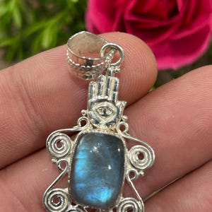 May include: A silver pendant featuring a rectangular labradorite gemstone with blue iridescence. The setting includes a hamsa hand symbol with an eye detail at the top and decorative silver wire swirls surrounding the central stone, finished with a bail.