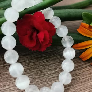 Pode incluir: Uma pulseira de contas brancas com contas redondas. Uma flor vermelha e folhas verdes estão no fundo. A pulseira é exibida em uma superfície de madeira.