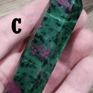 Op de afbeelding: Een groene en rode kristallen punt in een hand. Het kristal heeft een rechthoekige vorm met een puntige top. Het groen is bespikkeld met donkere vlekken en rode vlekken. De letter "C" is zichtbaar op het kristal.