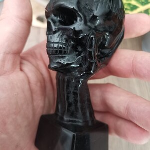 Peut inclure: Sculpture de crâne en obsidienne noire avec un visage détaillé et une base carrée. Le crâne est poli et a une finition brillante.