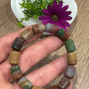Peut inclure: Un bracelet élastique composé de perles en pierre polie de forme cylindrique, déclinées dans des tons terreux variés comme le vert mousse, le jaune ocre, le brun profond et le gris sourd, assemblées sur un cordon flexible et uniforme.