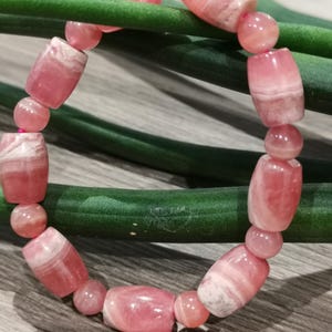 May include: A pink gemstone bracelet featuring a mix of barrel-shaped and round beads. The beads have a marbled appearance with varying shades of pink and white. The bracelet is displayed on a green stem.