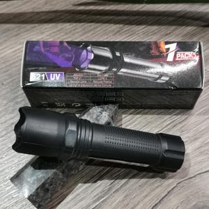 Könnte beinhalten: Eine schwarze UV-Taschenlampe mit strukturiertem Griff liegt auf einer grauen Oberfläche. Dahinter befindet sich eine schwarze Schachtel mit der Aufschrift "7 PACK". Die Schachtel trägt den Text "521 UV" und andere Texte.