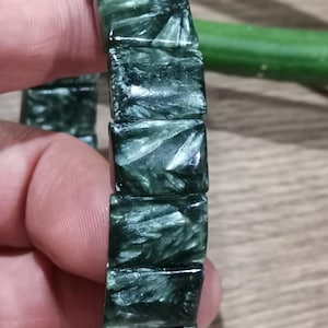 Peut inclure: Un bracelet vert foncé composé de pierres taillées en carrés. Les pierres présentent un aspect marbré avec des veines blanches et vert clair. Le bracelet est présenté sur un fond flou, mettant en valeur la texture et la couleur des pierres.