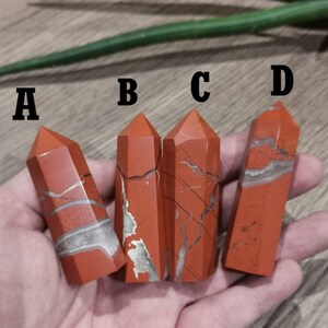 Peut inclure: Quatre tours en cristal de jaspe rouge avec des veines blanches et grises. Chaque tour a une forme et une taille différentes, étiquetées A, B, C et D. Les cristaux sont tenus dans une main.