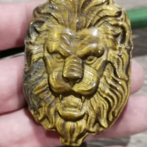 Op de afbeelding: Een gegraveerde hanger van tijgeroog in de vorm van een leeuwenkop. De steen is goud-bruin van kleur met bruine en gouden strepen.