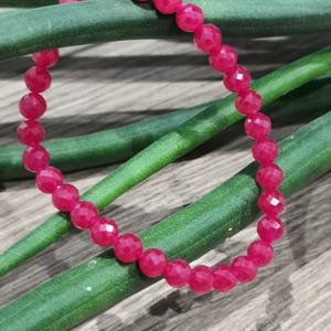 May include: A vibrant pink beaded bracelet with faceted beads. The bracelet is resting on a bed of green stems, with a wooden surface in the background. The beads are a deep, rich pink color.