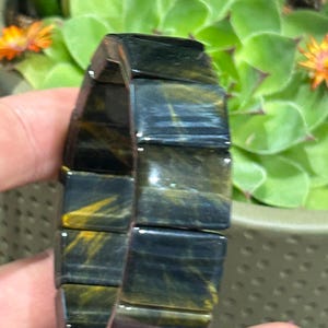 May include: A bracelet composed of rectangular, polished blue tiger's eye gemstone segments. Each stone piece features a chatoyant, fibrous texture with deep navy blue and golden-yellow streaks, linked together to form a continuous, flexible band.