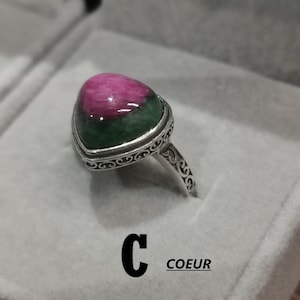 Op de afbeelding: Een zilveren ring met een groene en roze edelsteen. De ring heeft een decoratieve, sierlijke band. De tekst "C COEUR" is zichtbaar in de afbeelding.