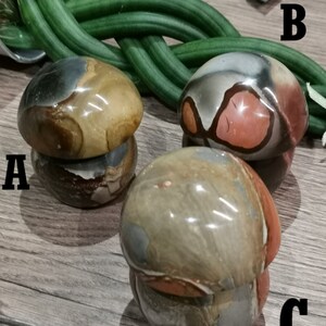 May include: Three mushroom-shaped polished stones with brown, gray, and red patterns. The stones are arranged on a wooden surface.
