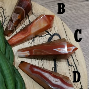 Pode incluir: Quatro varinhas de pedras preciosas polidas em tons de vermelho, laranja e branco são exibidas em uma superfície de madeira. Cada varinha tem uma ponta pontiaguda e um padrão único de faixas e inclusões. As varinhas são rotuladas A, B, C e D.