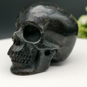 Peut inclure: Une sculpture de crâne en obsidienne noire. Le crâne est sculpté en détail, avec des orbites oculaires, un nez et des dents visibles.