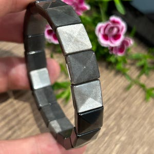 Könnte beinhalten: Ein elastisches Armband aus quadratischen, pyramidenförmigen Perlen in abwechselnden Farbtönen von metallischem Silber und mattem Schwarz. Die Perlen sind in einer durchgehenden Schlaufe angeordnet und weisen ein facettiertes Design auf.