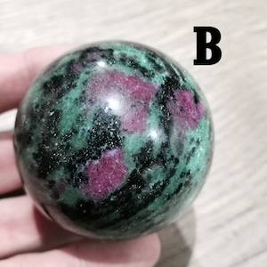 Puede incluir: Una esfera de piedra preciosa pulida. La esfera presenta una base verde oscuro con vetas negras y parches de color rojo intenso. La letra "B" es visible en el fondo.