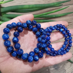 Könnte beinhalten: Drei blaue Perlenarmbänder mit poliertem Finish. Die Armbänder bestehen aus runden Perlen und werden in einer Hand gehalten. Der Hintergrund umfasst grüne Pflanzenstiele.