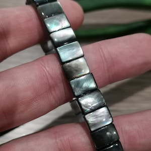 May include: A bracelet featuring rectangular links made of iridescent, grey-green mother-of-pearl. The bracelet is held in a hand, with a blurred green background. The links have a subtle sheen and are rectangular in shape.