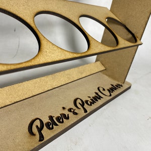 May include: Wooden paint rack with four circular cutouts for holding paint bottles. The rack has a brown stain and the words "Peter's Paint Center" are engraved on the front.
