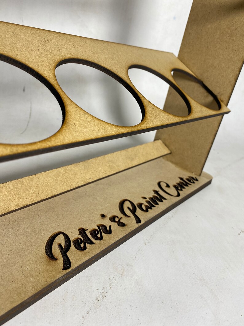 May include: Wooden paint rack with four circular cutouts for holding paint bottles. The rack has a brown stain and the words "Peter's Paint Center" are engraved on the front.