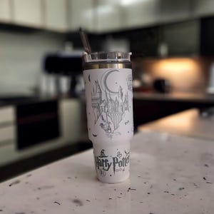 Puede incluir: Un vaso de acero inoxidable blanco con un gráfico en blanco y negro del castillo de Hogwarts y el texto "Harry Potter" en el lateral.
