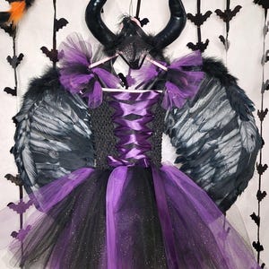 Puede incluir: Un tutú de tul negro y morado con alas y cuernos negros. El tutú tiene una cinta de satén morado alrededor de la cintura y purpurina negra por todas partes. Las alas son de plumas y los cuernos son negros.