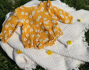 May include: A yellow baby onesie with a white daisy print, lying on a white crocheted blanket with fringe. The onesie has long sleeves and a ruffled neckline.