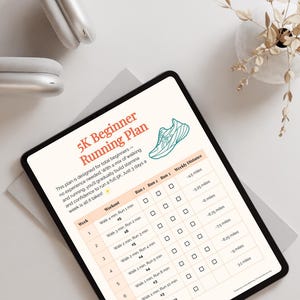May include: A digital tablet displays a 5K Beginner Running Plan with a running shoe graphic. The plan includes a weekly workout schedule with run and walk intervals, and weekly distance goals. A pair of white headphones and a small vase with dried plants are also visible.
