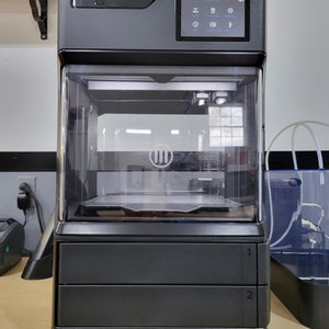 May include: A black and gray 3D printer with a clear plastic enclosure. The printer has a touchscreen display with the text "METHOD X" on the front. The printer is sitting on a wooden surface.