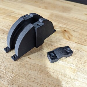 May include: Two black 3D printed parts, one is a curved bracket with two holes, the other is a smaller, curved clamp with two holes.