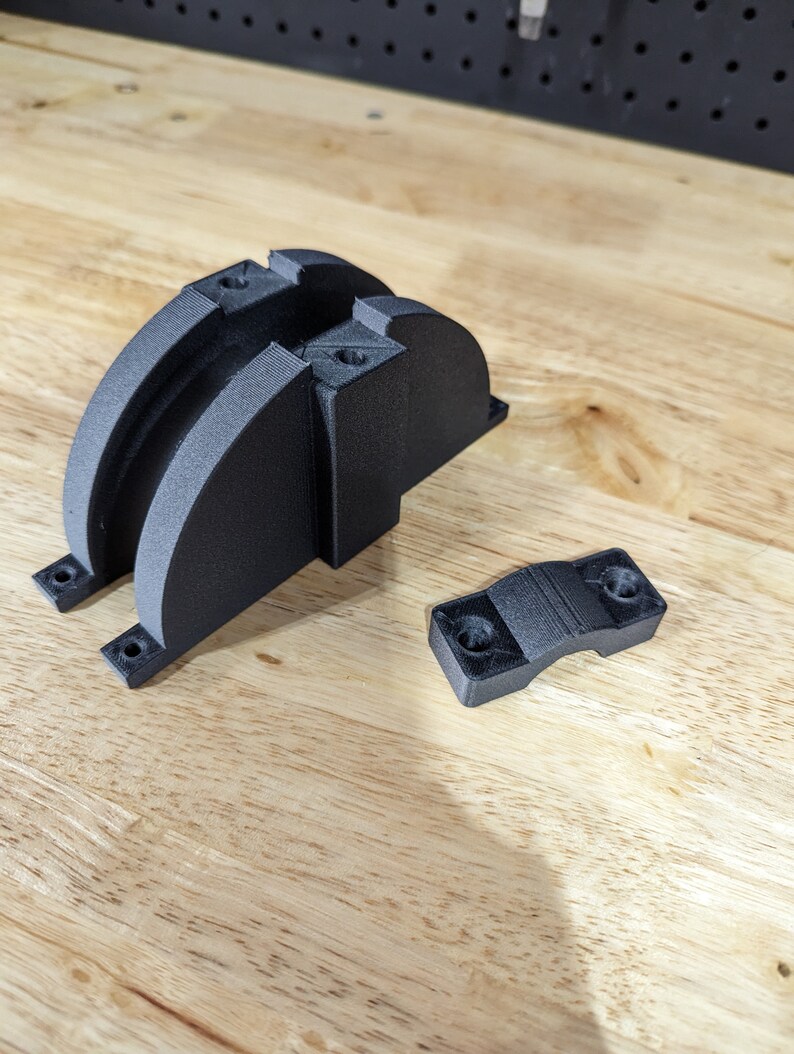 May include: Two black 3D printed parts, one is a curved bracket with two holes, the other is a smaller, curved clamp with two holes.