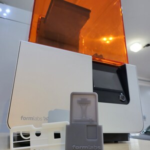 May include: A white 3D printer with an orange lid, a grey resin cartridge, and a white resin tray. The printer has the text "formlabs" on the front. The resin cartridge has the text "formlabs" on the front.