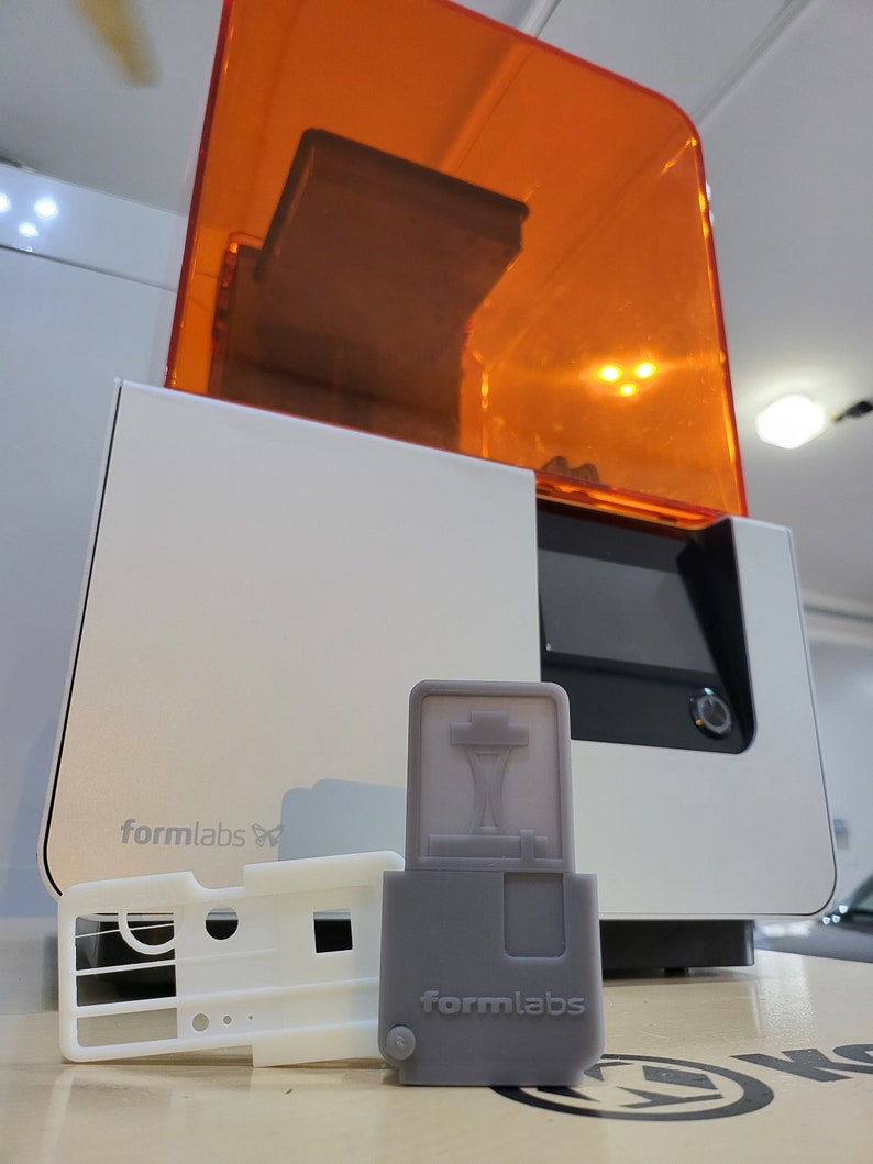 May include: A white 3D printer with an orange lid, a gray resin cartridge, and a white resin tray. The printer has the text "formlabs" on the front. The resin cartridge has the text "formlabs" on the front.