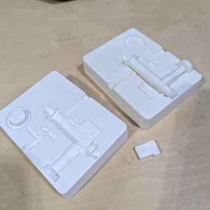 May include: Two white plastic molds for casting a small metal pump. The molds are open and show the details of the pump design.