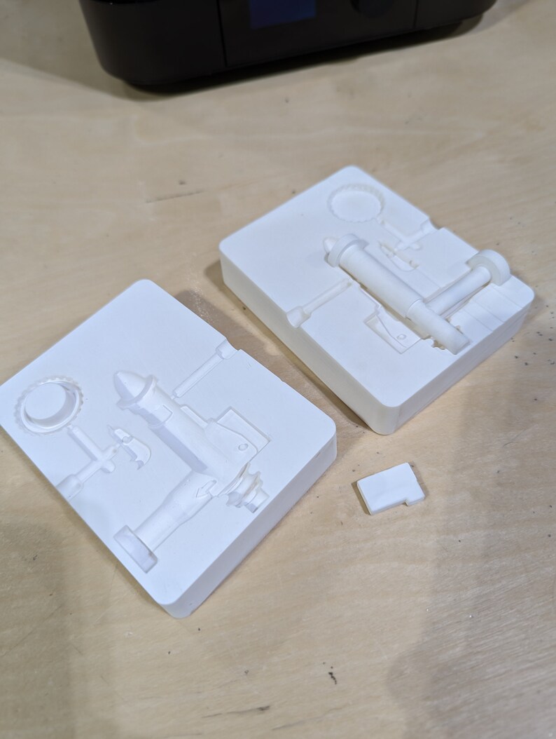 May include: Two white plastic molds for casting a small metal pump. The molds are open and show the details of the pump design.