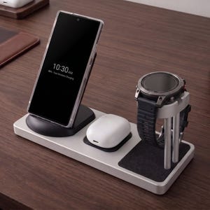 May include: A silver wireless charging station with a phone, smartwatch, and earbuds. The phone displays 10:30 AM and is charging. The watch has a black strap. The earbuds are white.