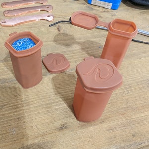 May include: Three brown silicone containers with lids. The containers are cylindrical and have a wave design on the lids. One container has a blue lid with a white design.