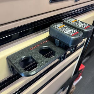May include: Black plastic battery holder with the text "Overly Engineered" printed on it. The holder is attached to a beige metal toolbox drawer. Two black Snap-on batteries are in the holder.