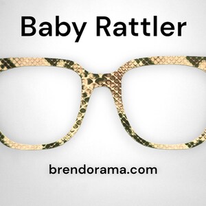 May include: A pair of brown and green patterned spectacles with a square frame. The glasses have a snakeskin pattern. The text "Baby Rattler" is above the glasses and "brendorama.com" is below the glasses.