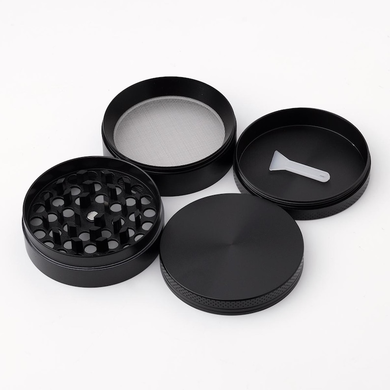 May include: A four-piece black metal grinder with a screen and a scraper. The grinder has a textured surface and a smooth, flat top.