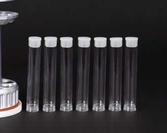 May include: Seven clear, cylindrical vials with white caps are arranged in a row against a black background. The vials are made of transparent plastic, allowing a view of the contents. The caps are flat and opaque.