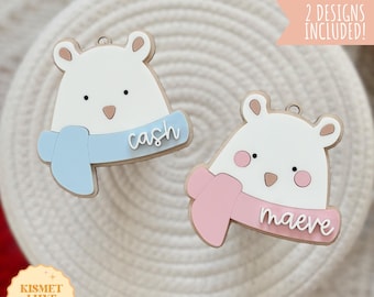 May include: Two laser-cut wooden ornaments shaped like polar bears wearing scarves. The ornament on the left has a blue scarf and the text "cash" on it. The ornament on the right has a pink scarf and the text "maeve" on it.