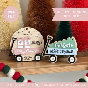 May include: Two wooden Christmas ornaments shaped like a wagon. The first wagon is decorated with a pink gift box, a pink banner that says "Merry Christmas", and the name "Aubrey". The second wagon is decorated with a green Christmas tree, a blue banner that says "Merry Christmas", and the name "mason". Both wagons have white wheels and a black frame.