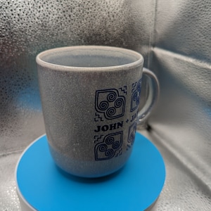 May include: A gray ceramic mug with a blue design featuring the text "JOHN + JAN" and a swirling pattern. The mug is sitting on a blue turntable.